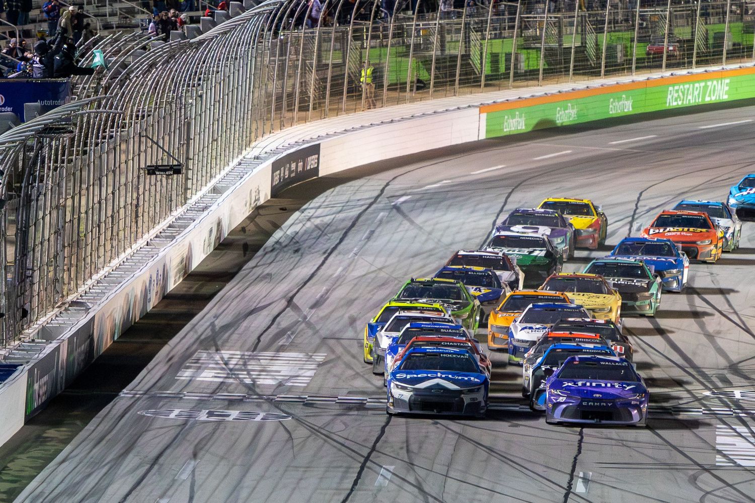 NASCAR race cars compete closely on an oval track during a night race at EchoPark Speedway