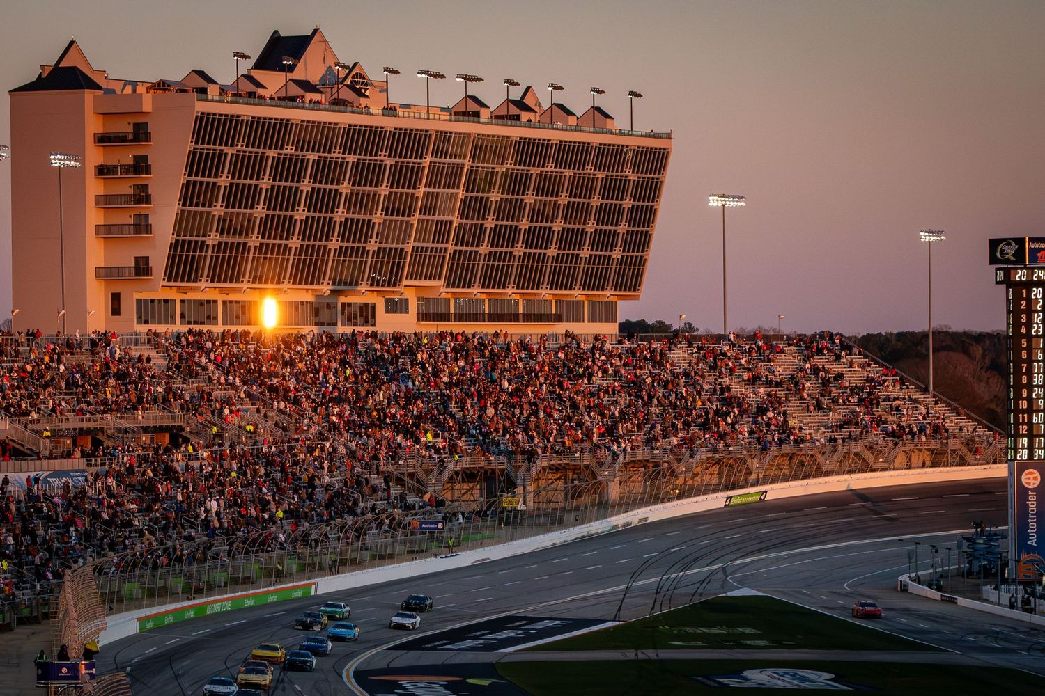 Packed grandstands at NASCAR track during sunset with stadium lights illuminated.