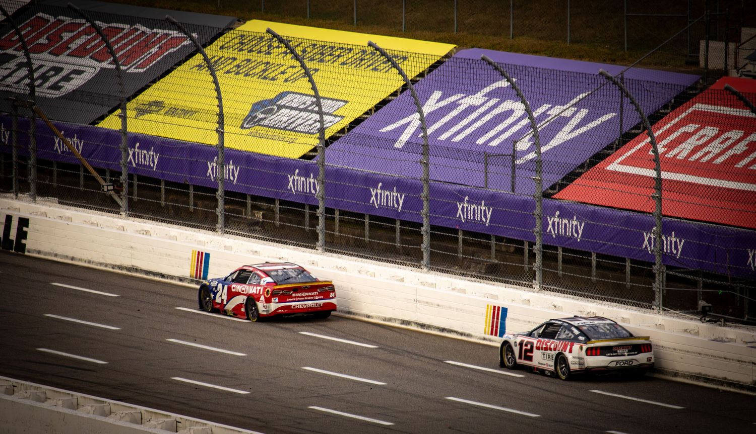 NASCAR vehicles race past yellow and purple track markings during a competitive event.
