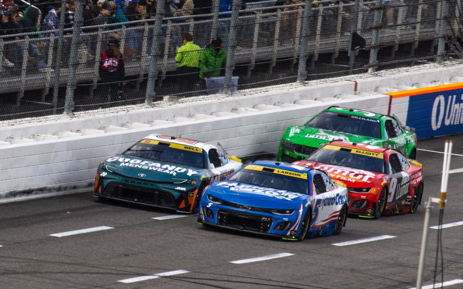 NASCAR race cars competing on an oval track with grandstands visible in the background.