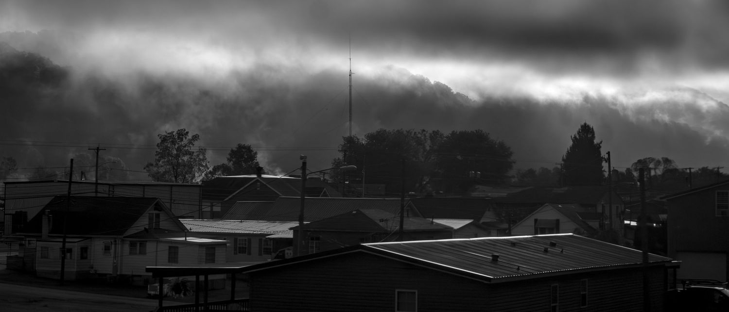 Moody black and white cityscape with dark storm clouds looming over industrial buildings and rooftops.