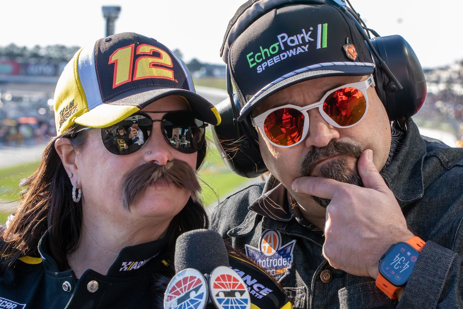 Two racing enthusiasts with matching mustaches pose together wearing sunglasses and NASCAR apparel.
