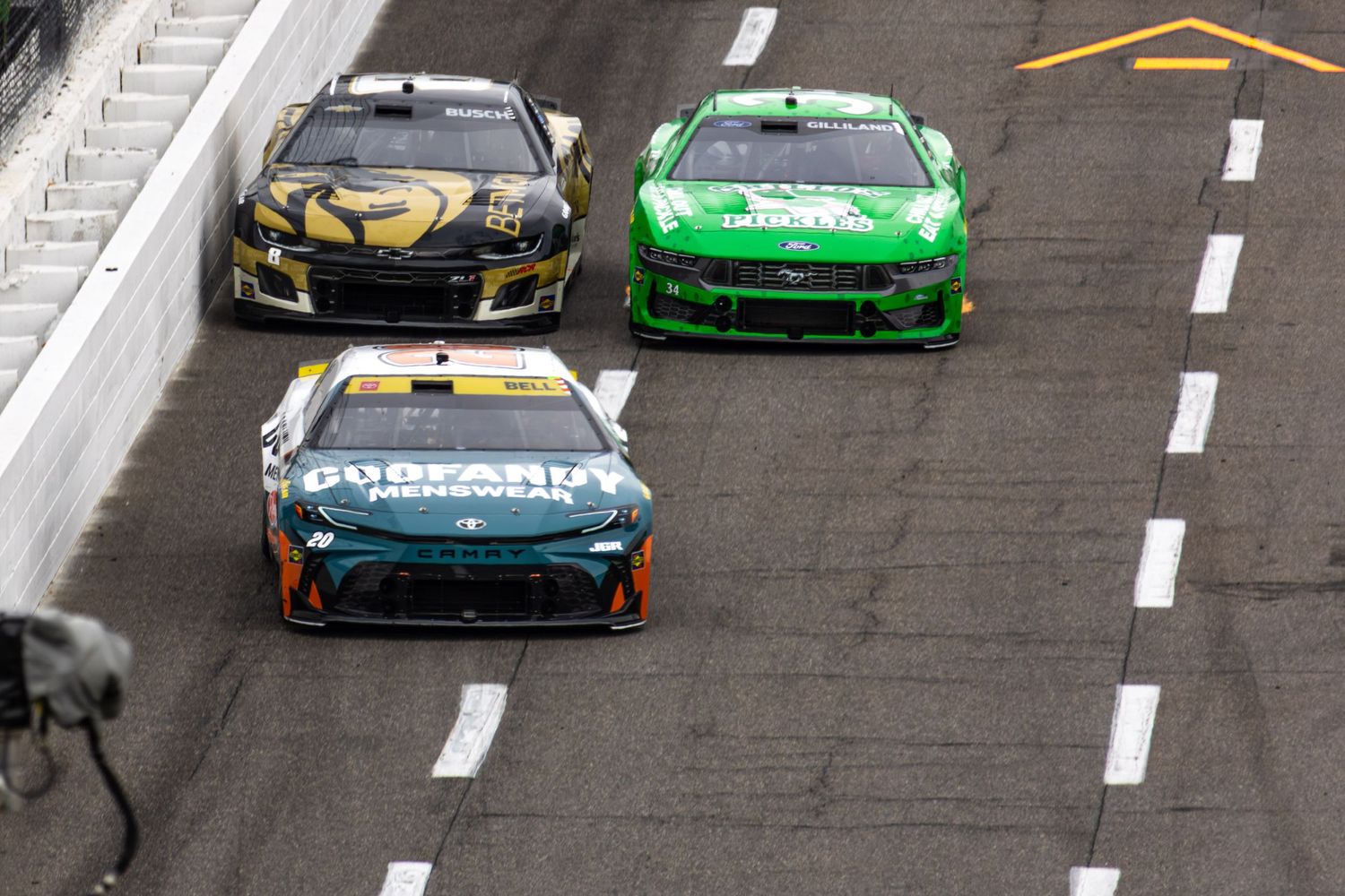 NASCAR race cars compete on an oval track with yellow and white markings visible on the asphalt surface.