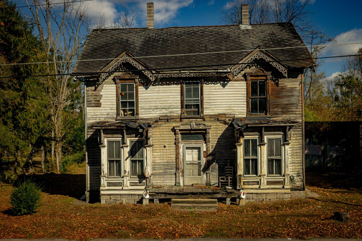 Panoramic view of a weathered white farmhouse with dormers and wrap-around porch in autumn setting.
