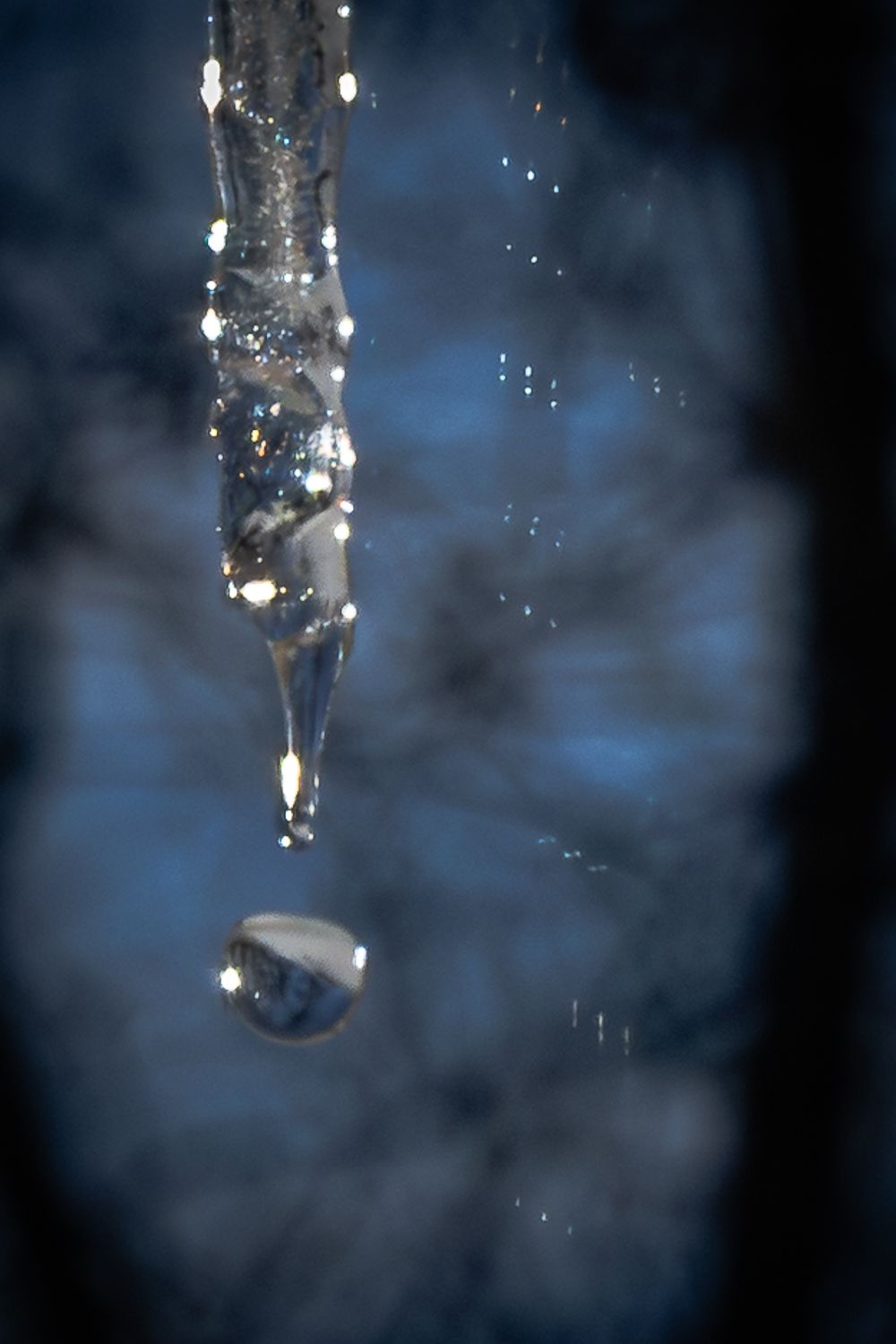 A frozen icicle drips a single water droplet against a blurred blue background.