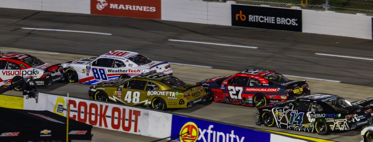 NASCAR race cars compete closely together during a nighttime race at a speedway with Cook Out and Xfinity sponsorship visible.