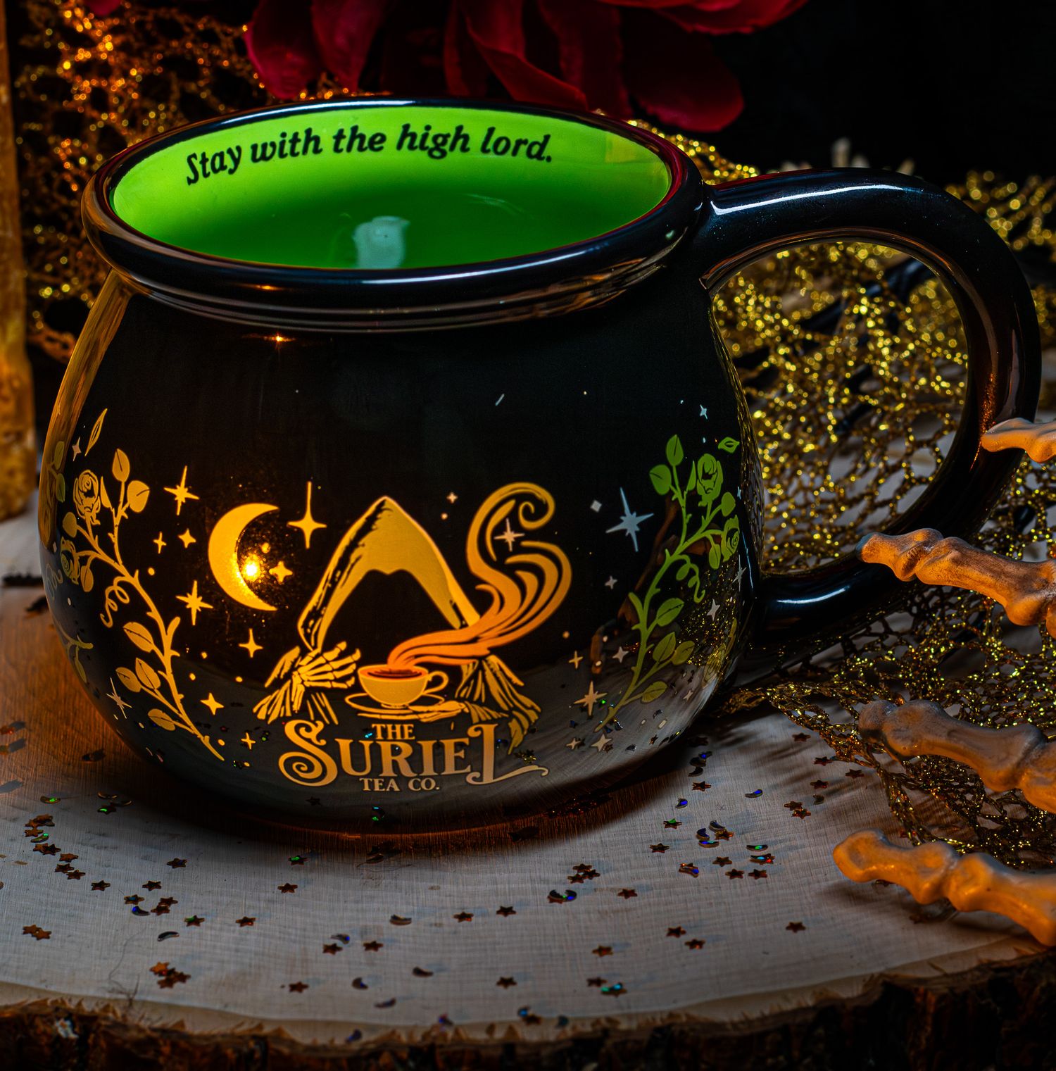 Black mug with gold Suriel design and green interior, reading Stay with the high lord, on a wooden surface.