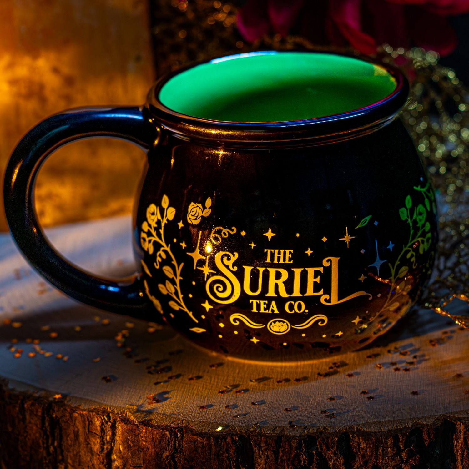 Black and green ceramic mug with gold The Suriel Tea Co. design, placed on a wooden surface with warm ambient lighting.