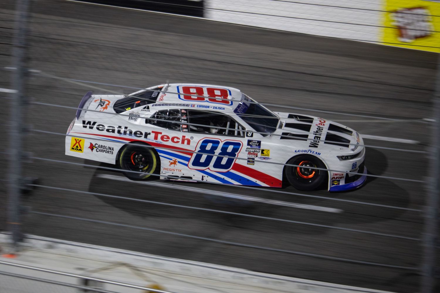 White WeatherTech NASCAR race car speeds past on oval track during competition.