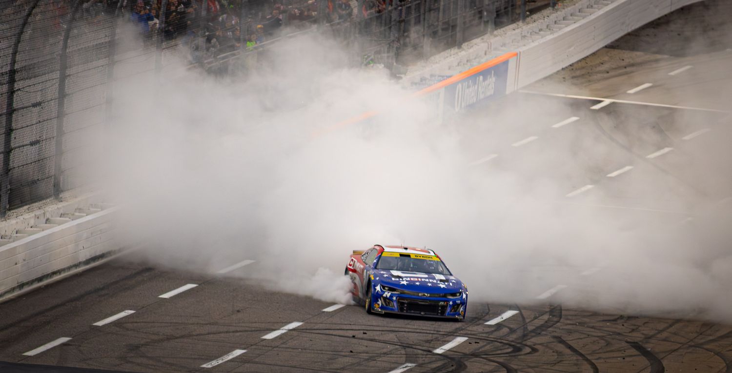 Blue stock car creates dramatic smoke cloud during victory celebration burnout.