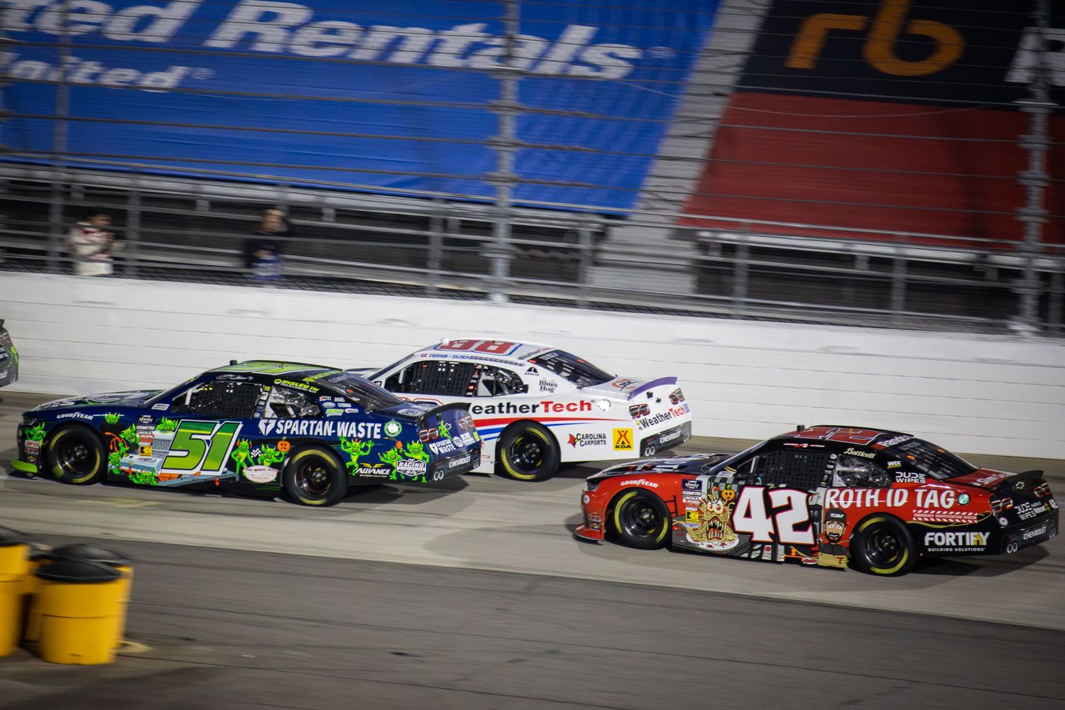 Multiple NASCAR vehicles race closely together on an oval track during competition.