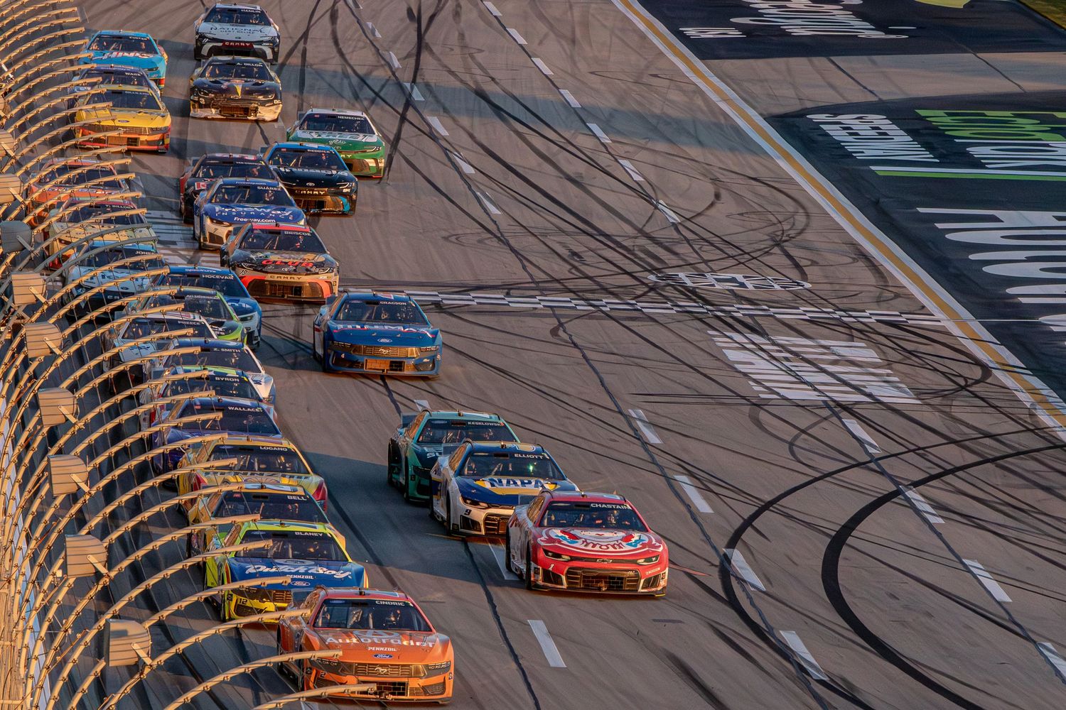 NASCAR race cars navigate turn in tight pack formation leaving tire marks on track.