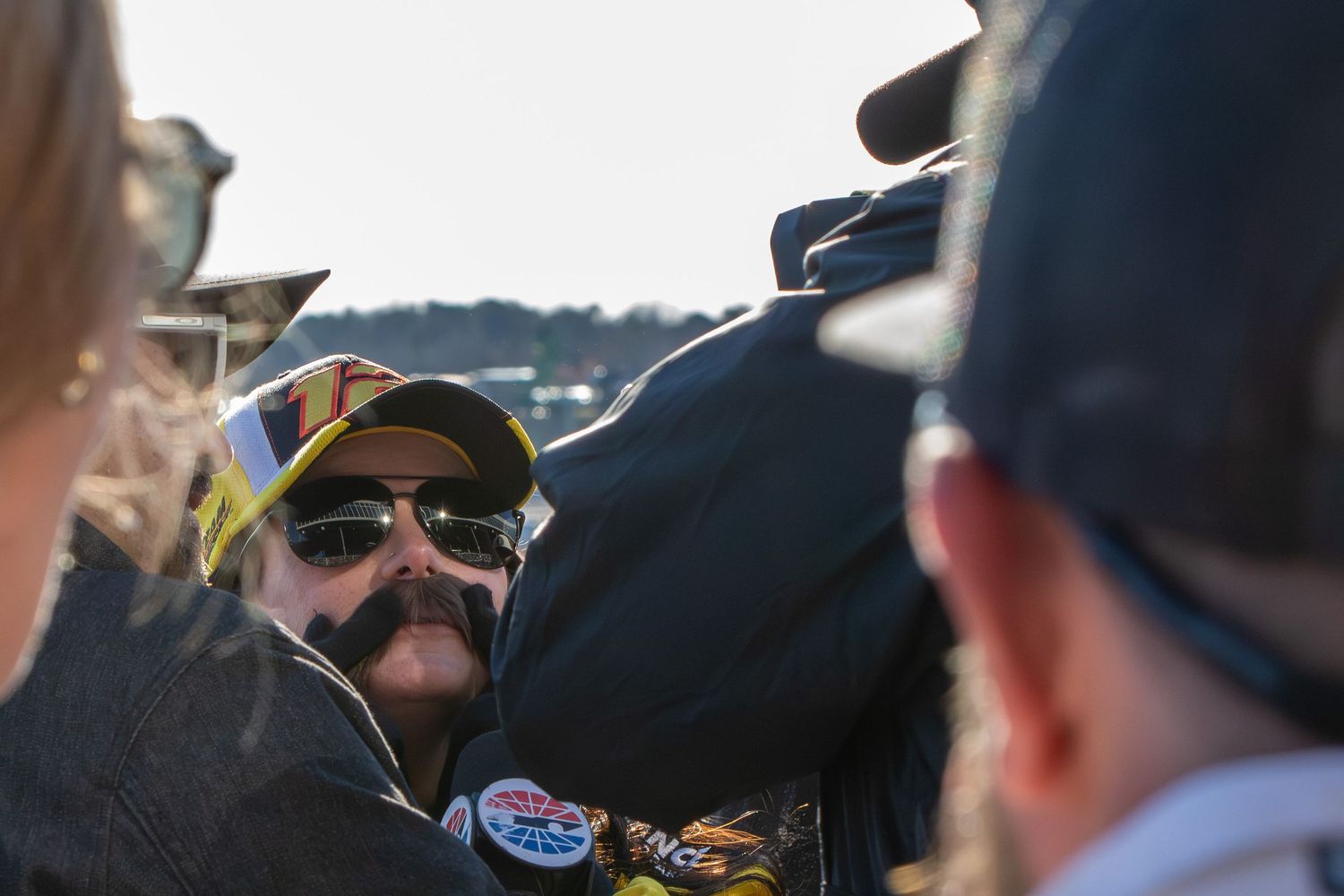 Racing fans gather at a NASCAR event wearing team merchandise and sunglasses on a sunny day.