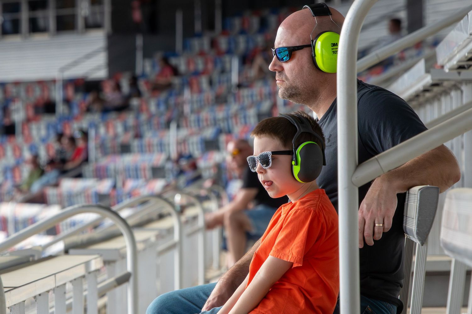 Spectators wearing protective headphones watch intently from stadium seats during a motorsports event.
