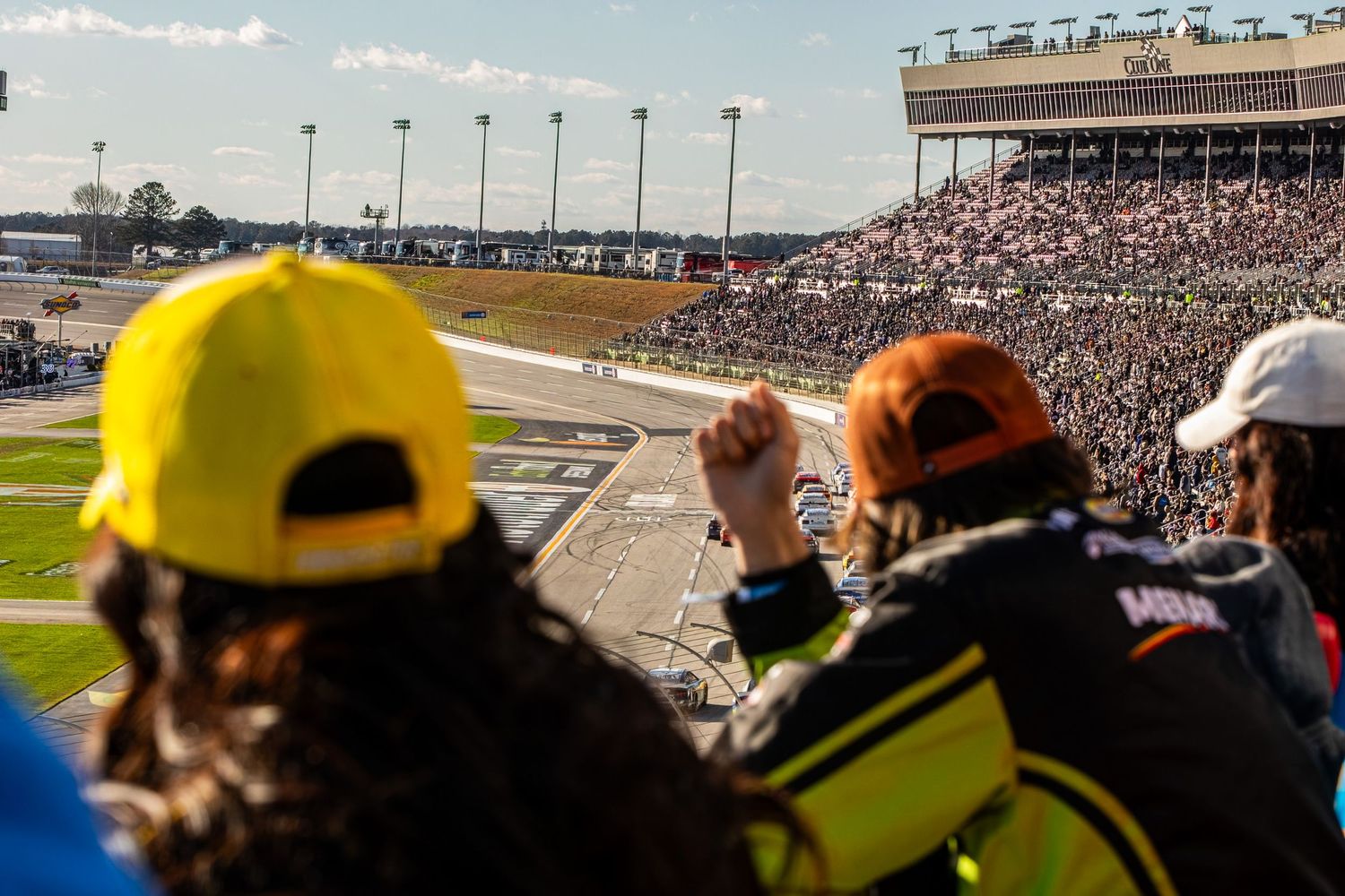 Crowded grandstand of racing fans watching cars race on an oval track under bright sunlight.