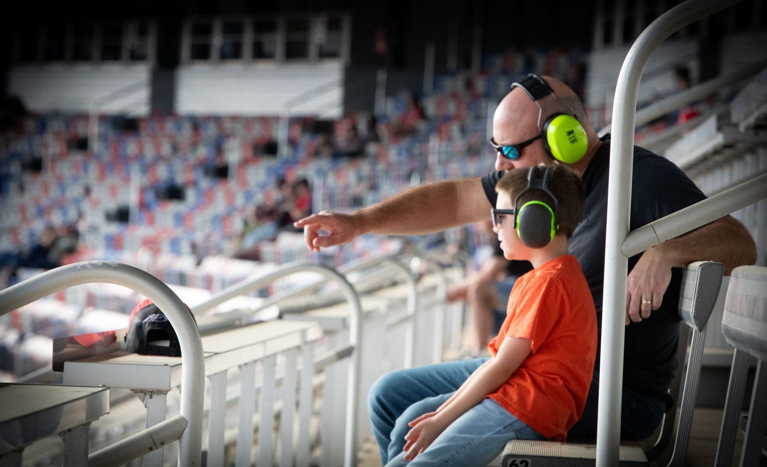 Young spectator in bright orange shirt wearing ear protection watches racing action from stadium seating.
