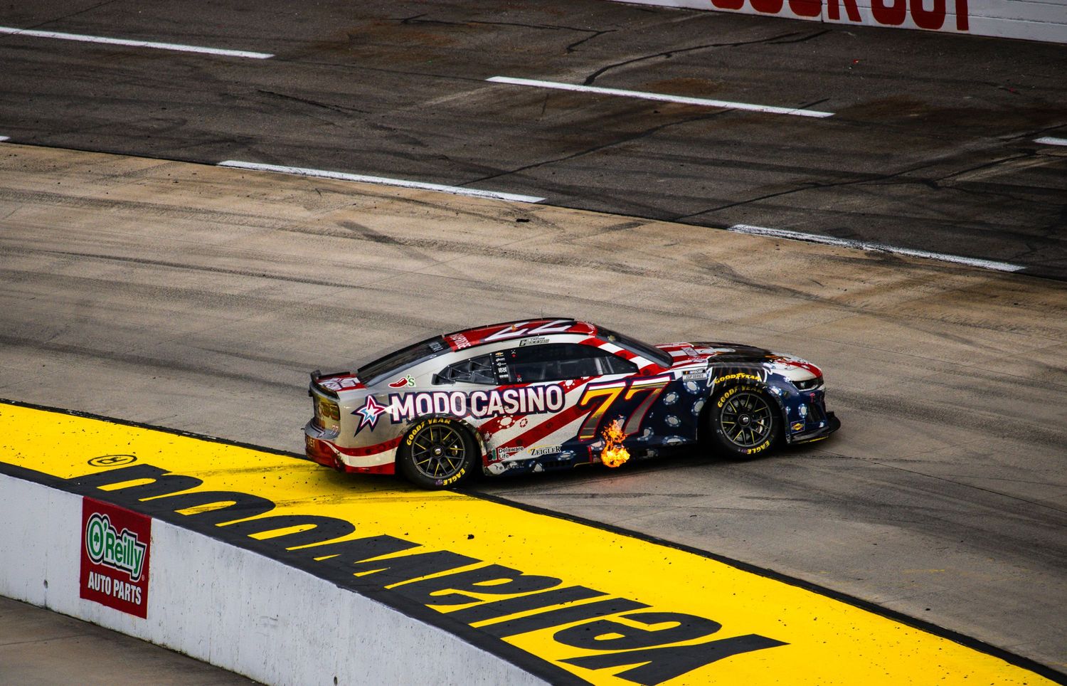 Race cars speed past sponsorship logos painted on the track surface during a NASCAR competition.