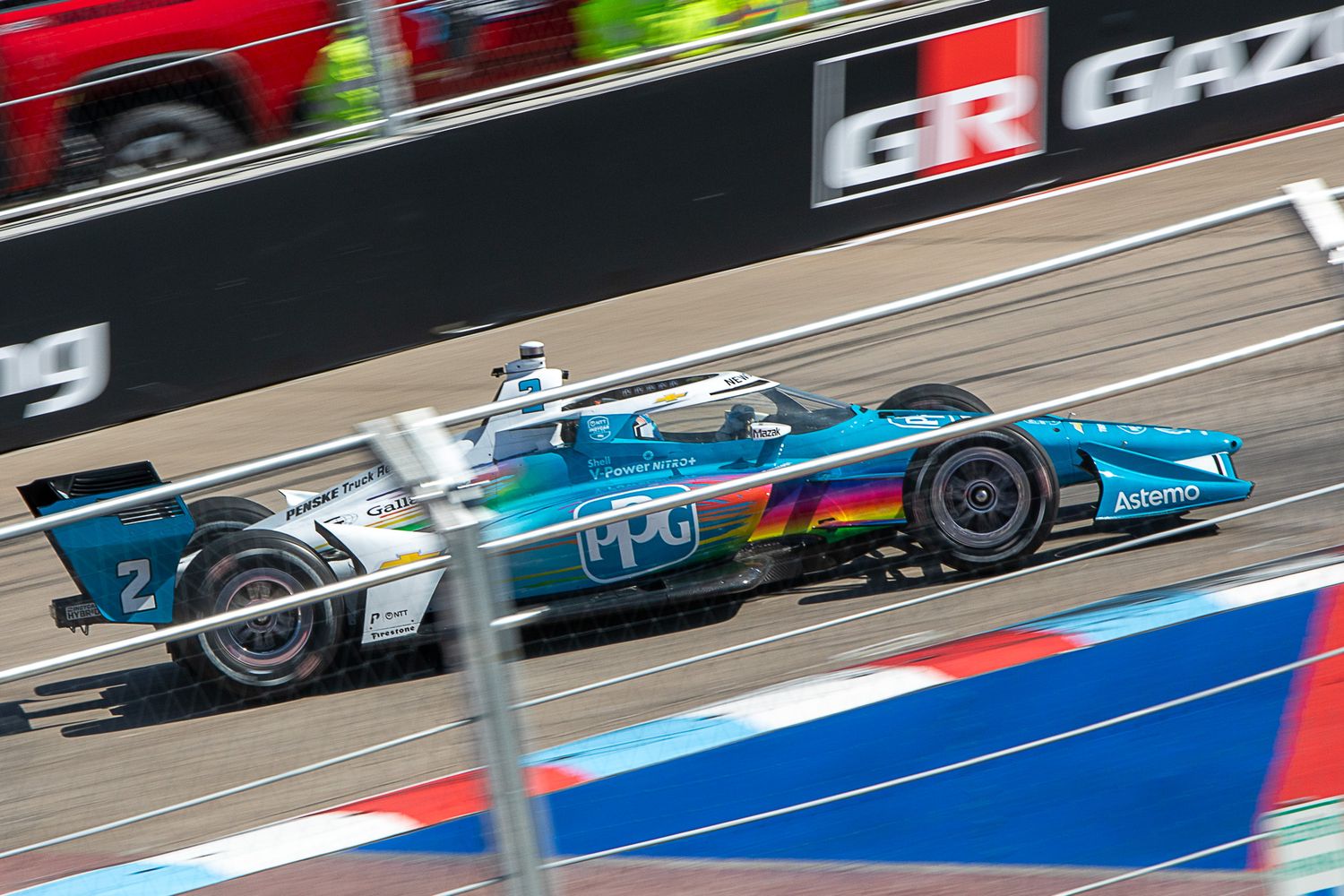 Formula E electric race car speeds past on racetrack with blue and red barriers visible in background.