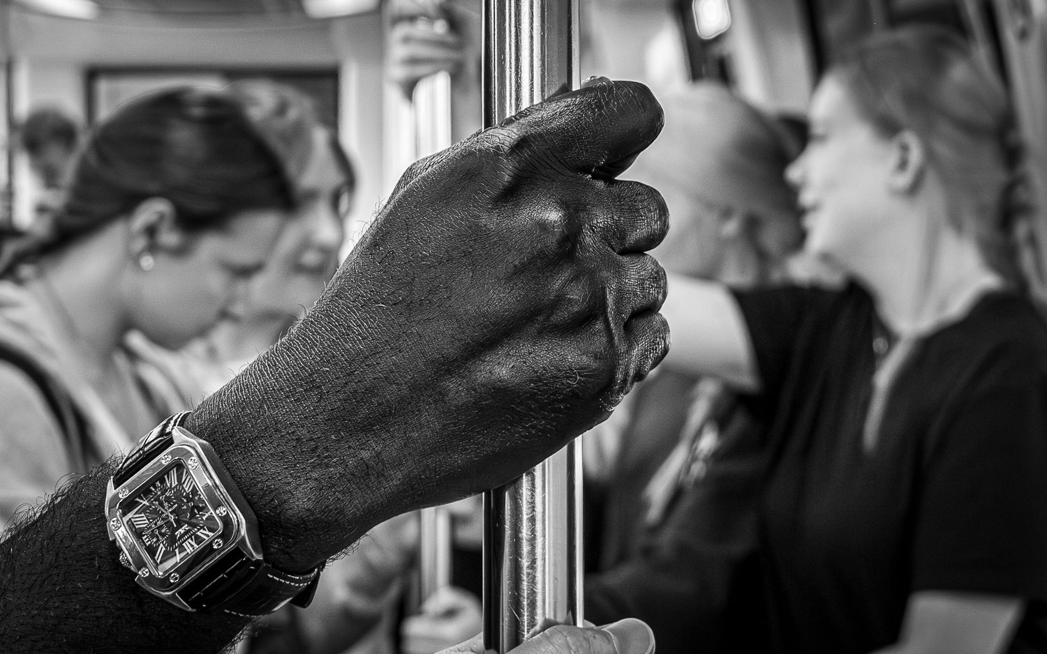 Black and white photo of a man's hand gripping a subway pole, wearing a watch, with passengers in the background.