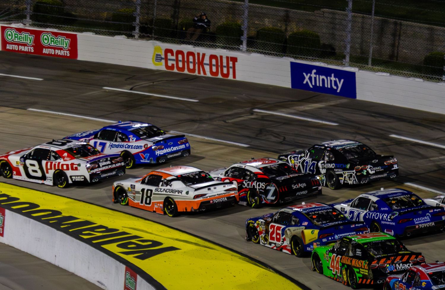 Stock cars race in tight formation on an illuminated NASCAR track with Cook Out and Xfinity advertising boards.