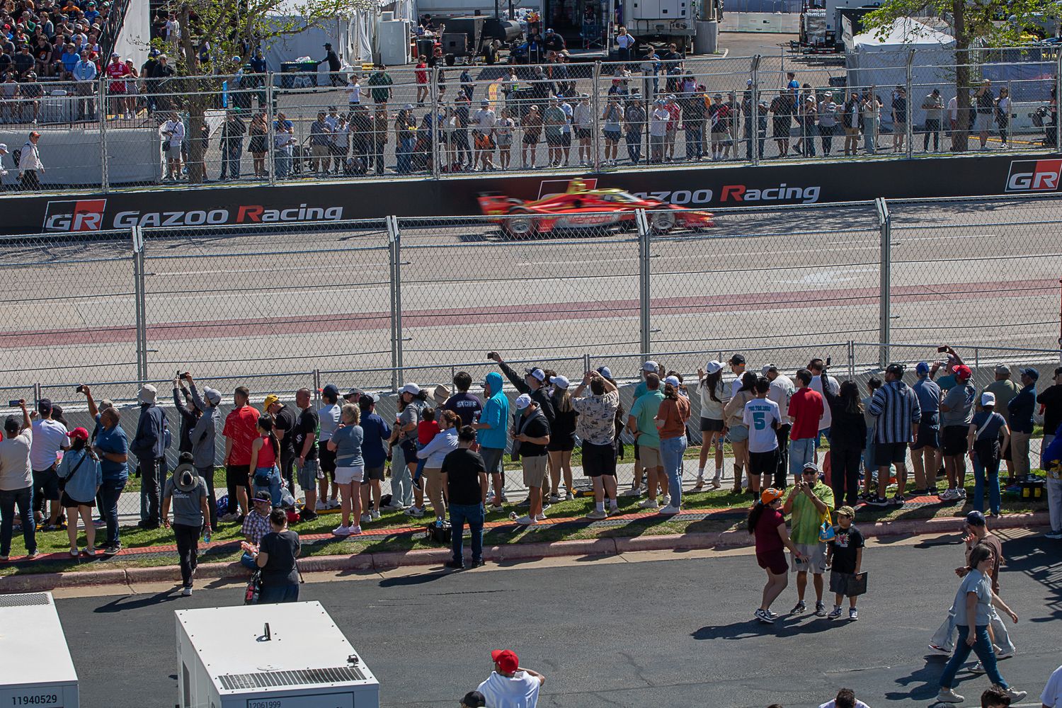 Spectators line the fence at a motorsport race track watching race cars speed past on a sunny day.