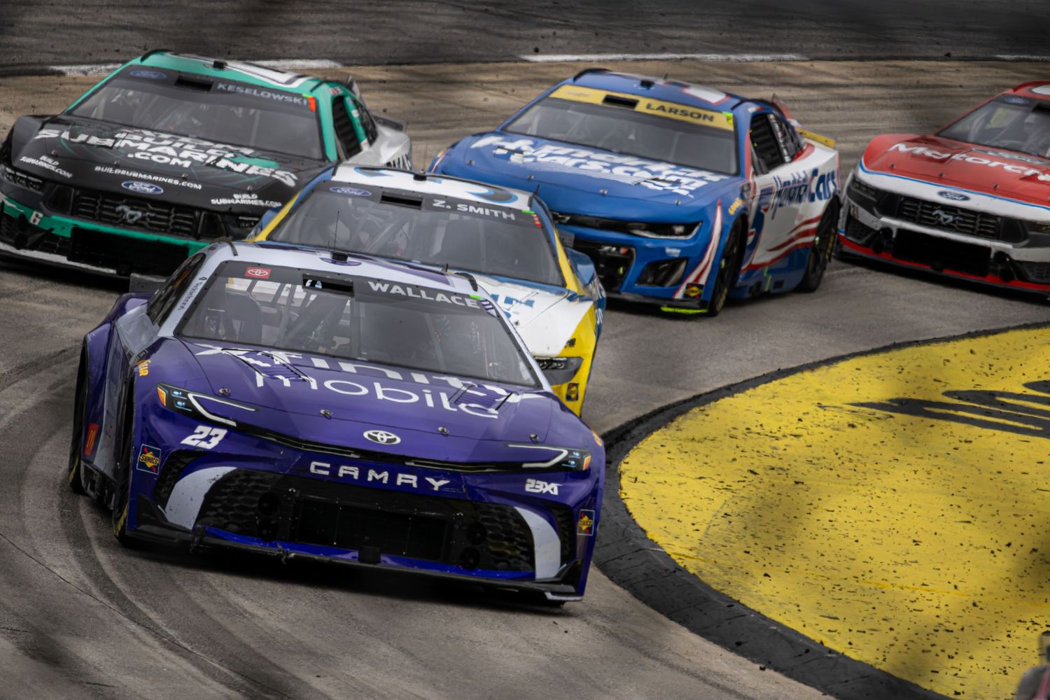 Multiple stock cars racing closely together on a NASCAR track with colorful sponsor liveries.