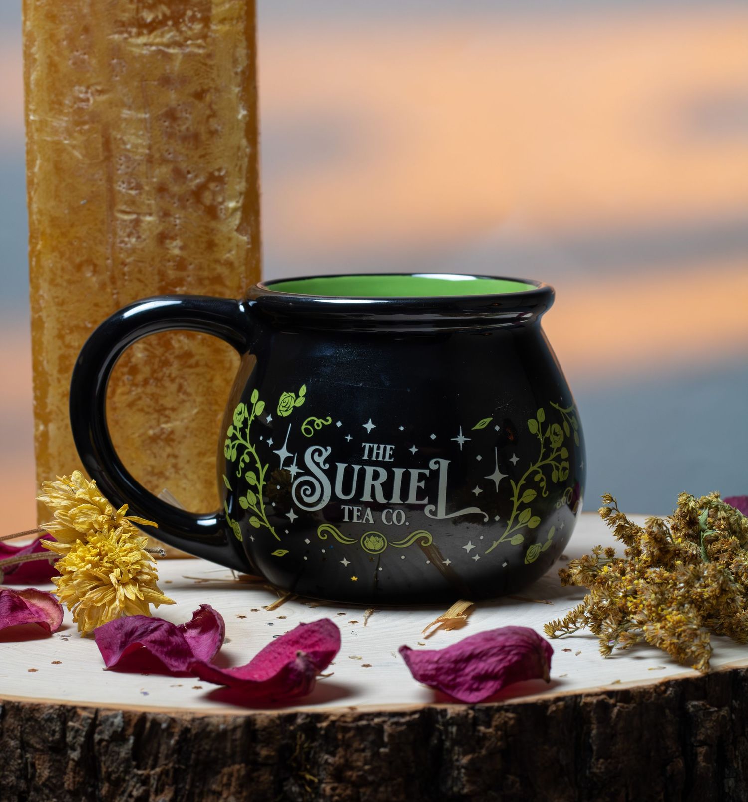 Black cauldron-shaped mug with green Suriel Tea Co. logo, surrounded by dried flowers on a wood slice at sunset.