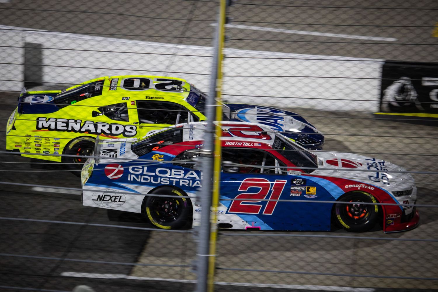 NASCAR race cars compete side by side on track with bright yellow Menards car battling against red and blue competitors.
