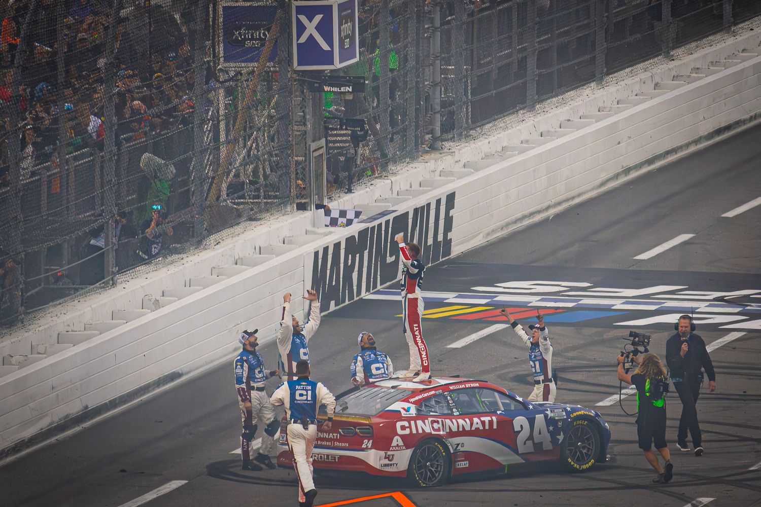 Emergency crews assess damage to wrecked NASCAR vehicle on race track.