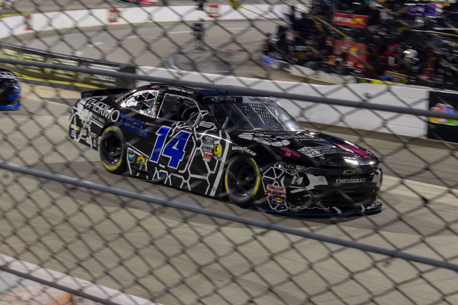 Dark NASCAR stock car with number 14 races past chain link fence during night race.