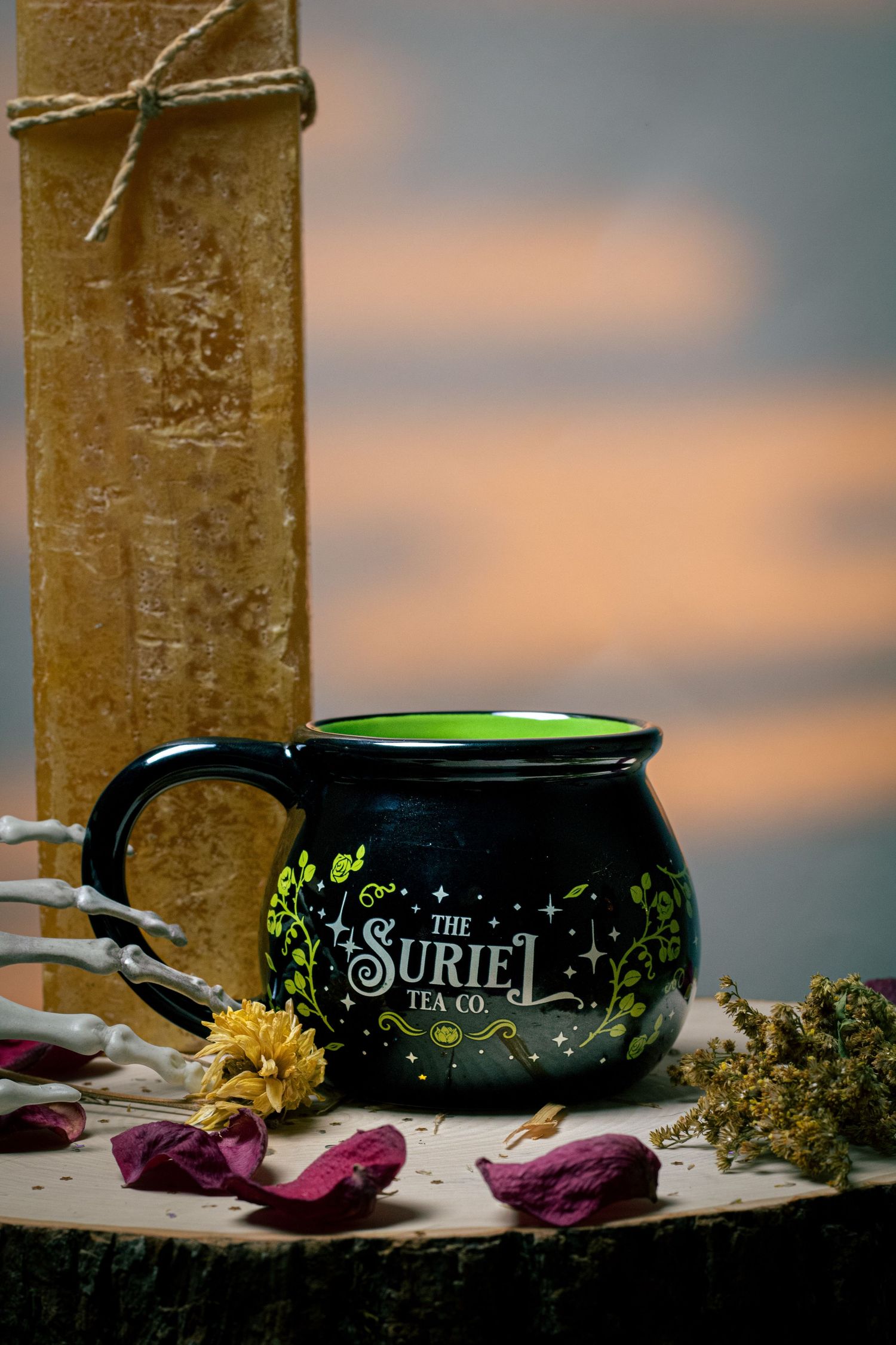Black cauldron-shaped mug with green interior and The Suriel Tea Co. logo, surrounded by dried flowers and skeleton hand.