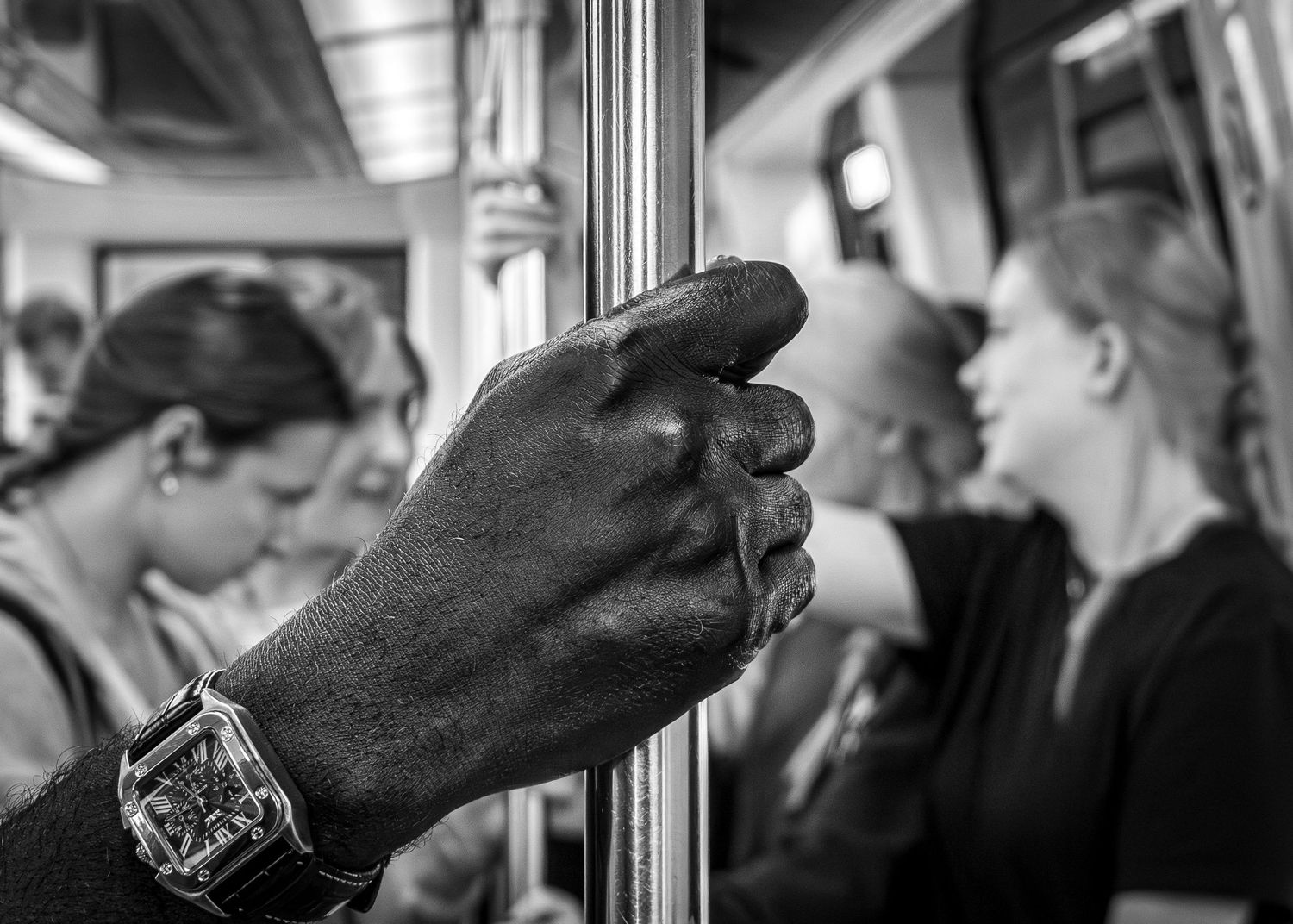 Close-up of a hand gripping a subway pole, wearing a watch, with blurred passengers in the background.