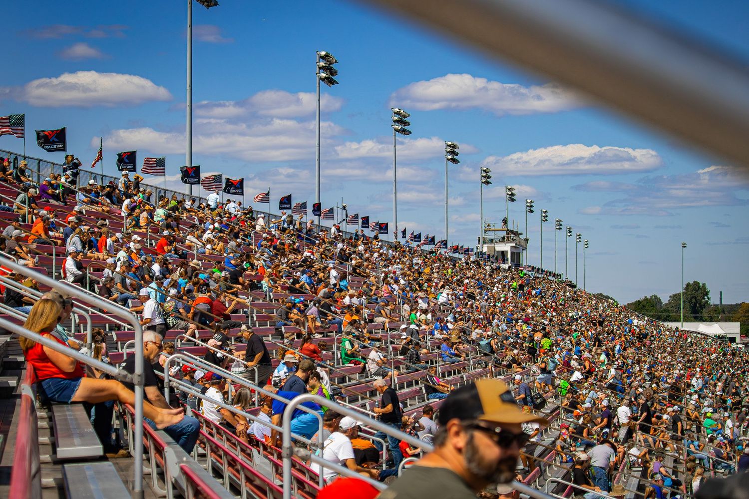 Packed grandstands filled with racing fans at a NASCAR event under blue skies.