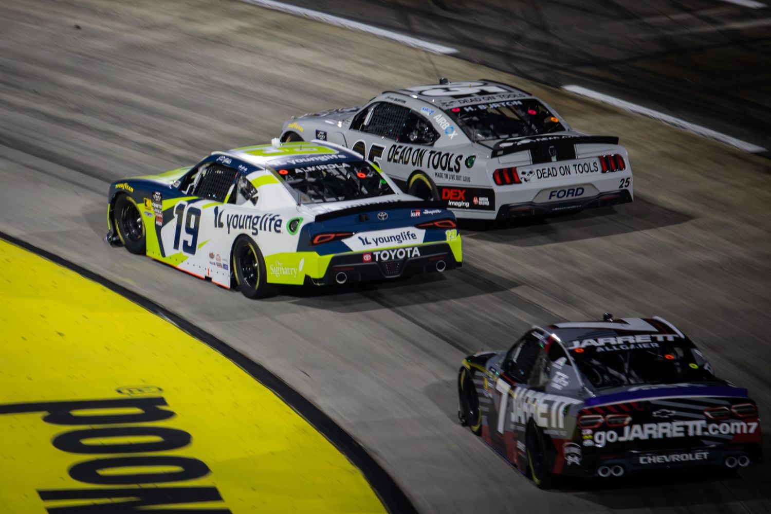Race cars navigate a turn with yellow track markings under nighttime lighting conditions.
