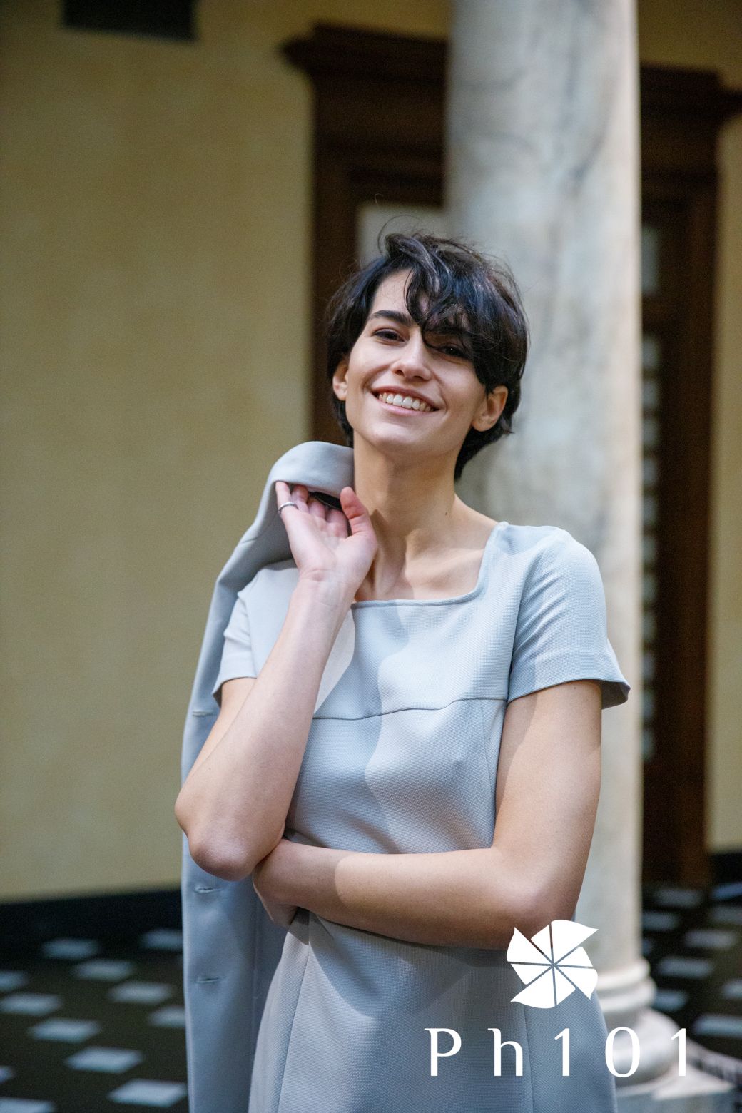 Figure in casual grey attire poses playfully against a beige wall with columns.