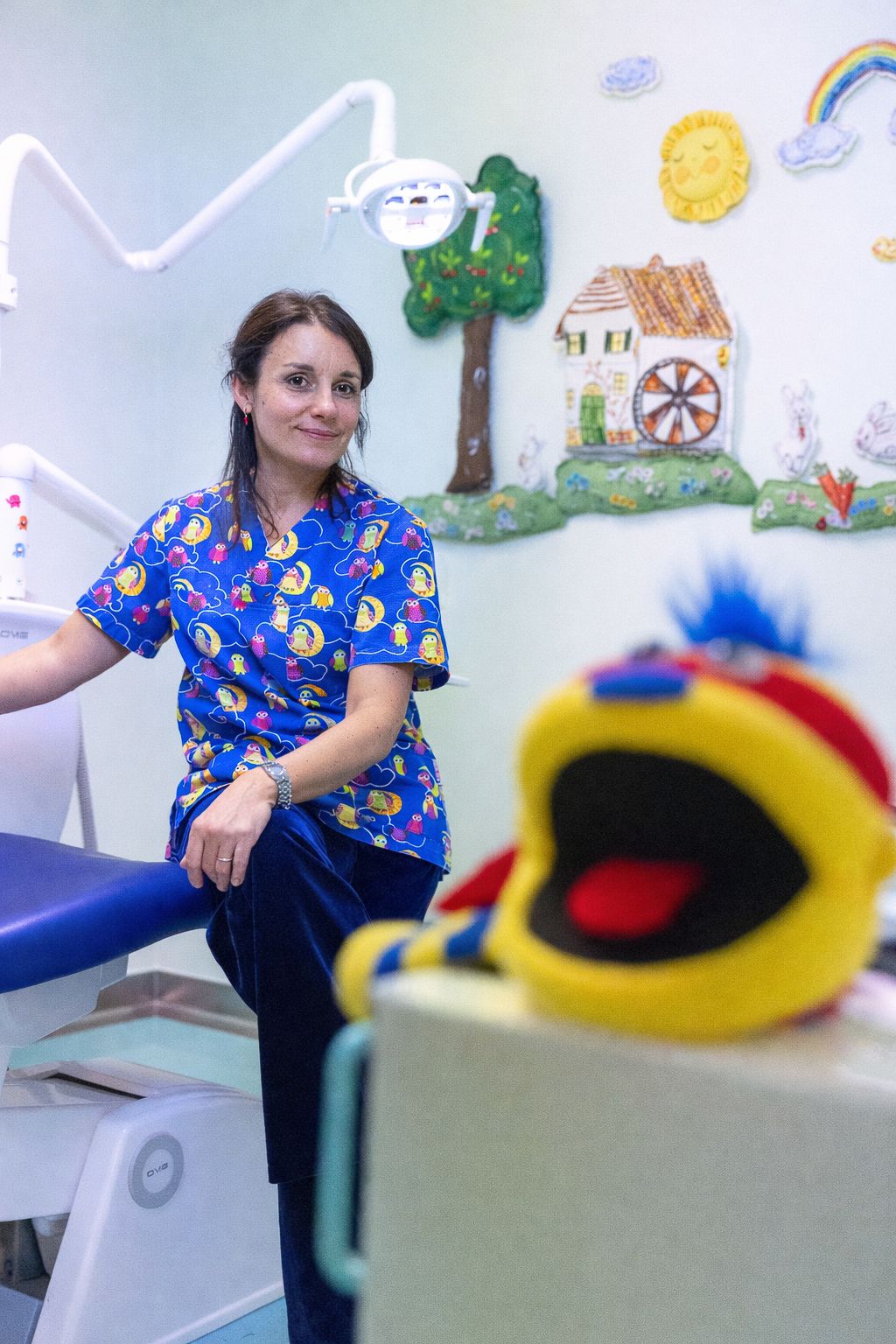 A person in a blue patterned shirt sits in a classroom with colorful wall art and a plush toy in the foreground.