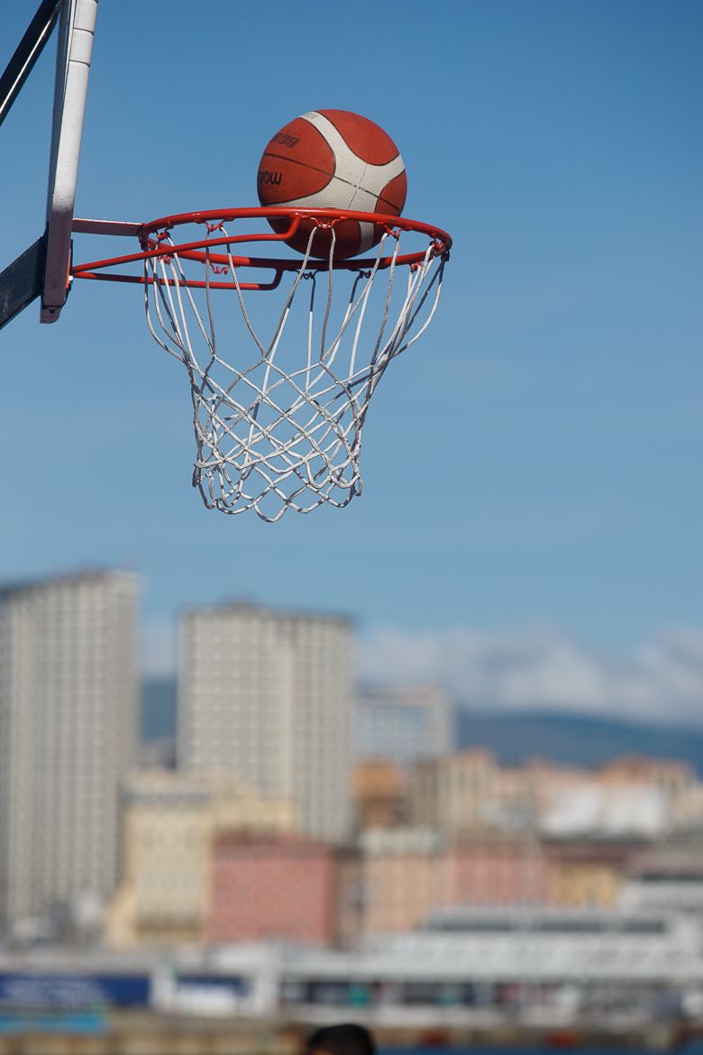 Basketball balanced on rim against blue sky with city buildings in background.