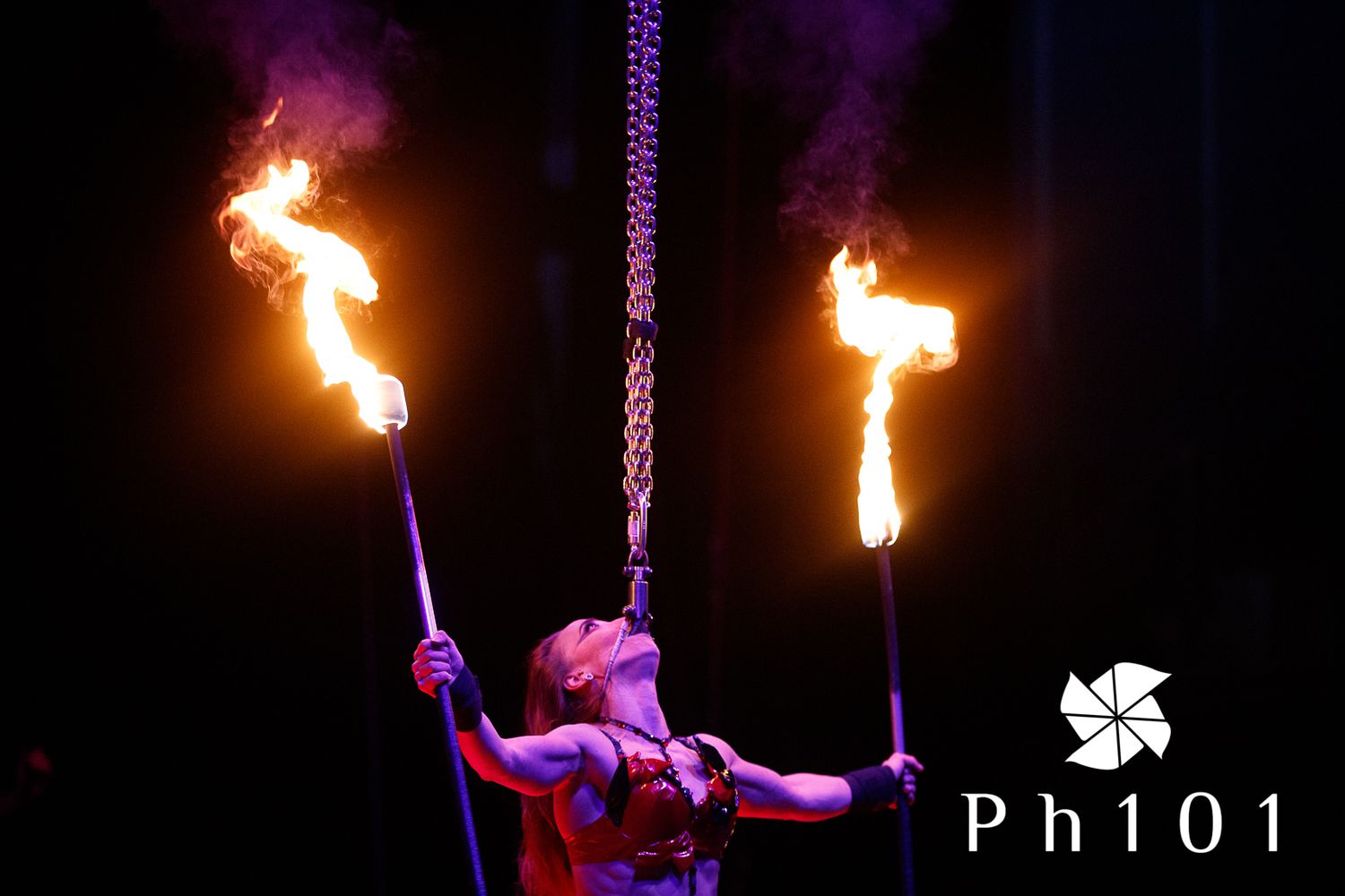 Fire performer twirling flaming batons against a dark background with purple lighting.