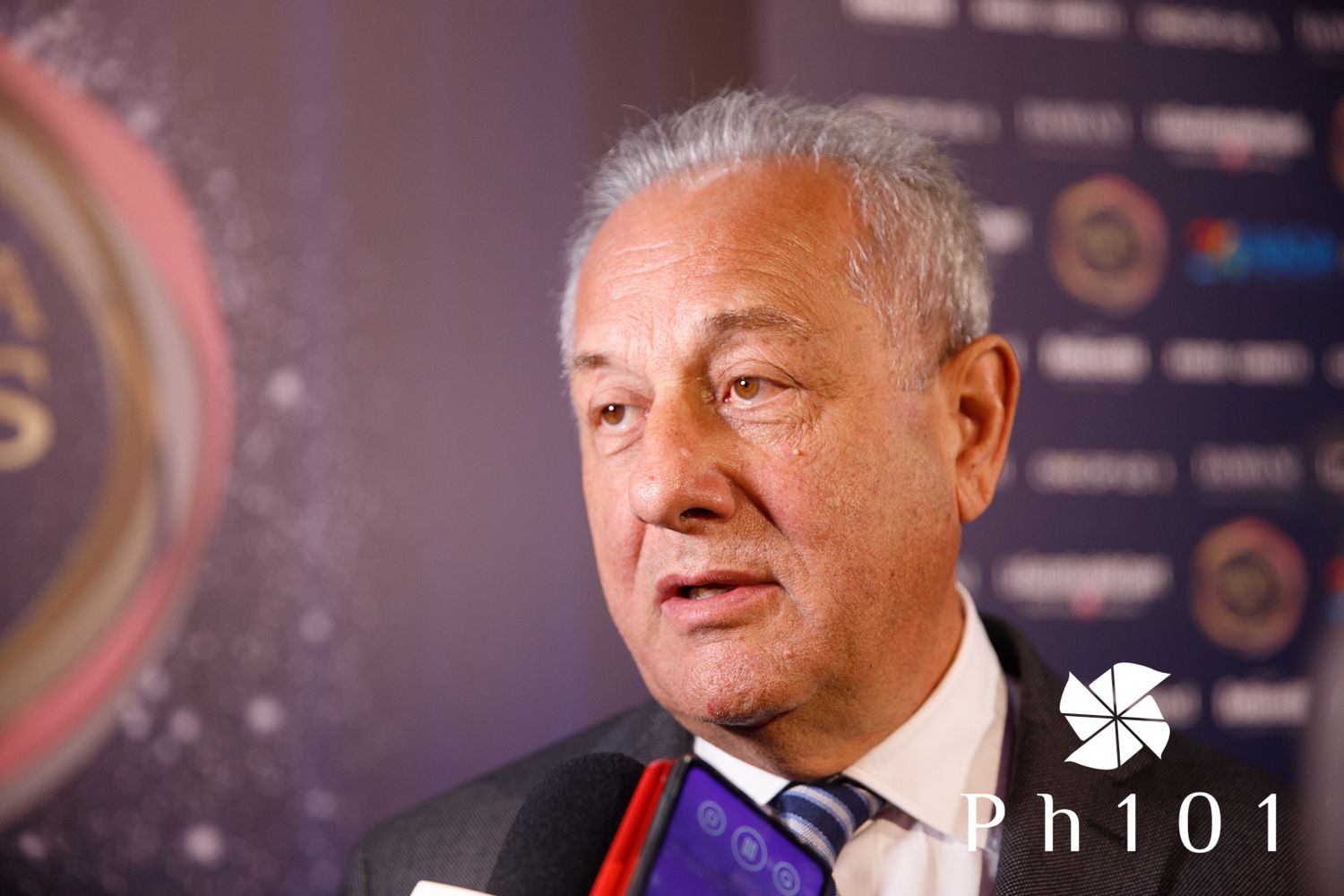 A speaker wearing formal attire with a medal at what appears to be a press event.