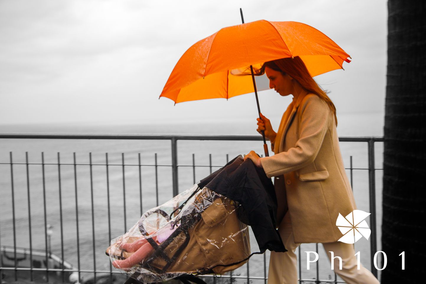 Person in beige coat holding an orange umbrella while sitting near a waterfront on an overcast day.