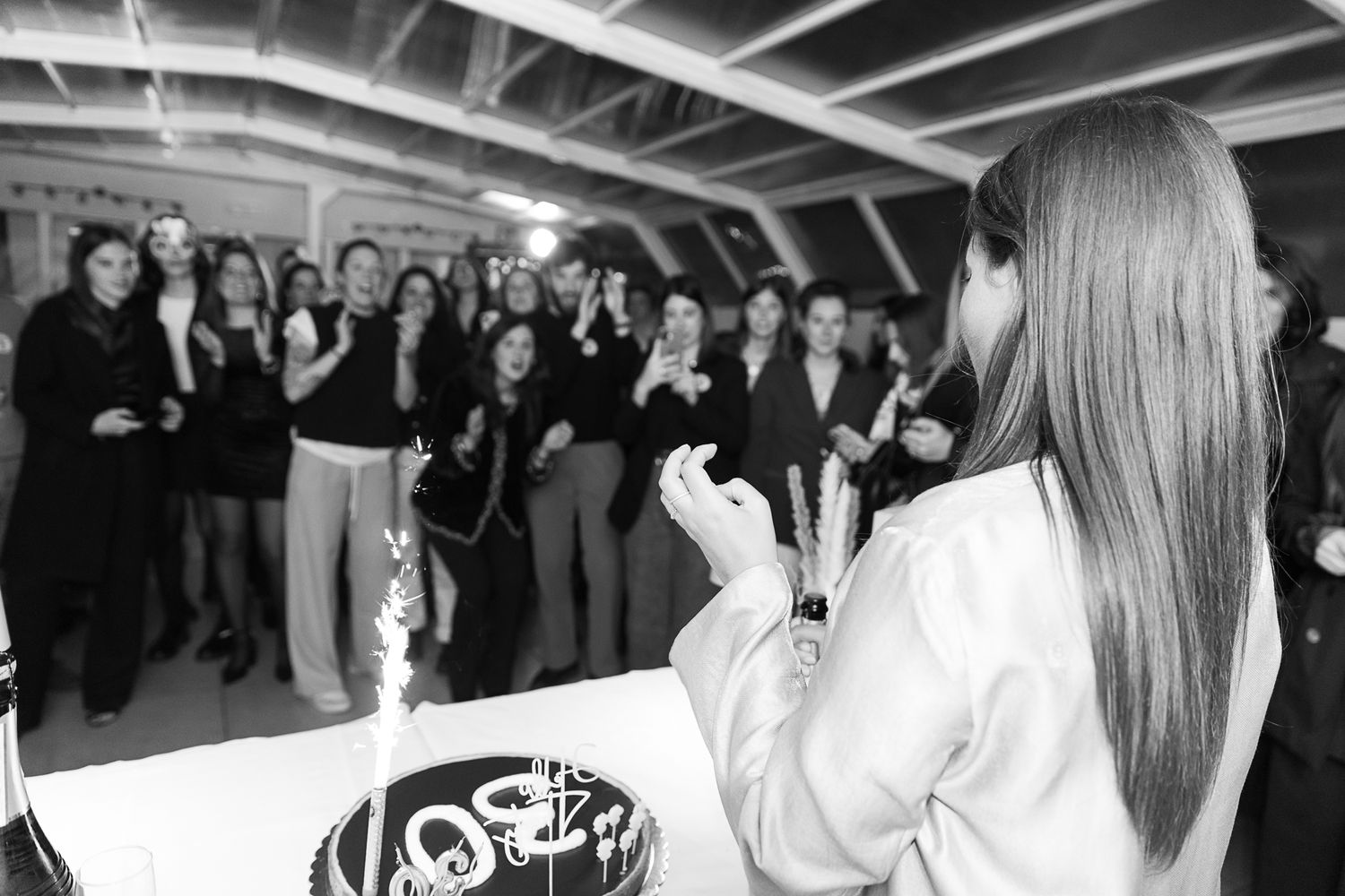 Black and white photo of a woman cutting a birthday cake while a group of people clap and celebrate in the background.