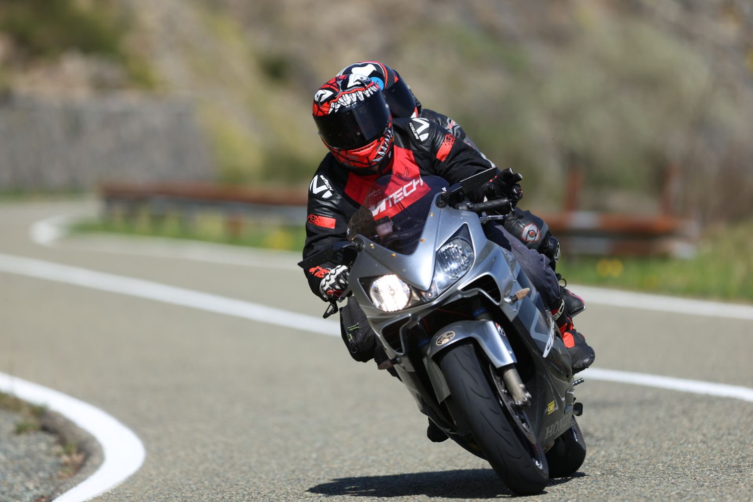 Motorcyclist in red gear riding a sport bike through a winding mountain road on a sunny day.