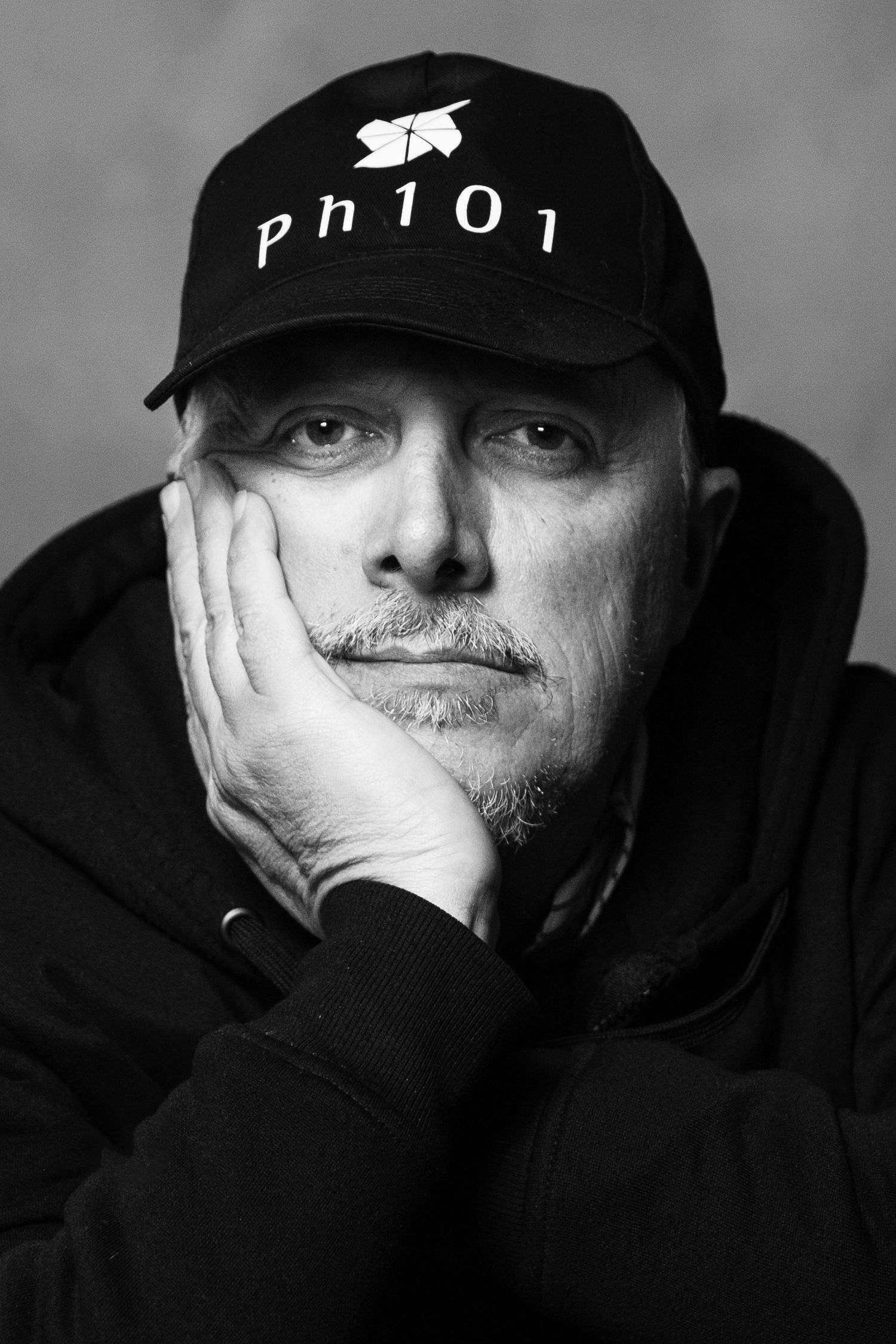 Moody portrait of person in black cap and hoodie with hand resting on face.