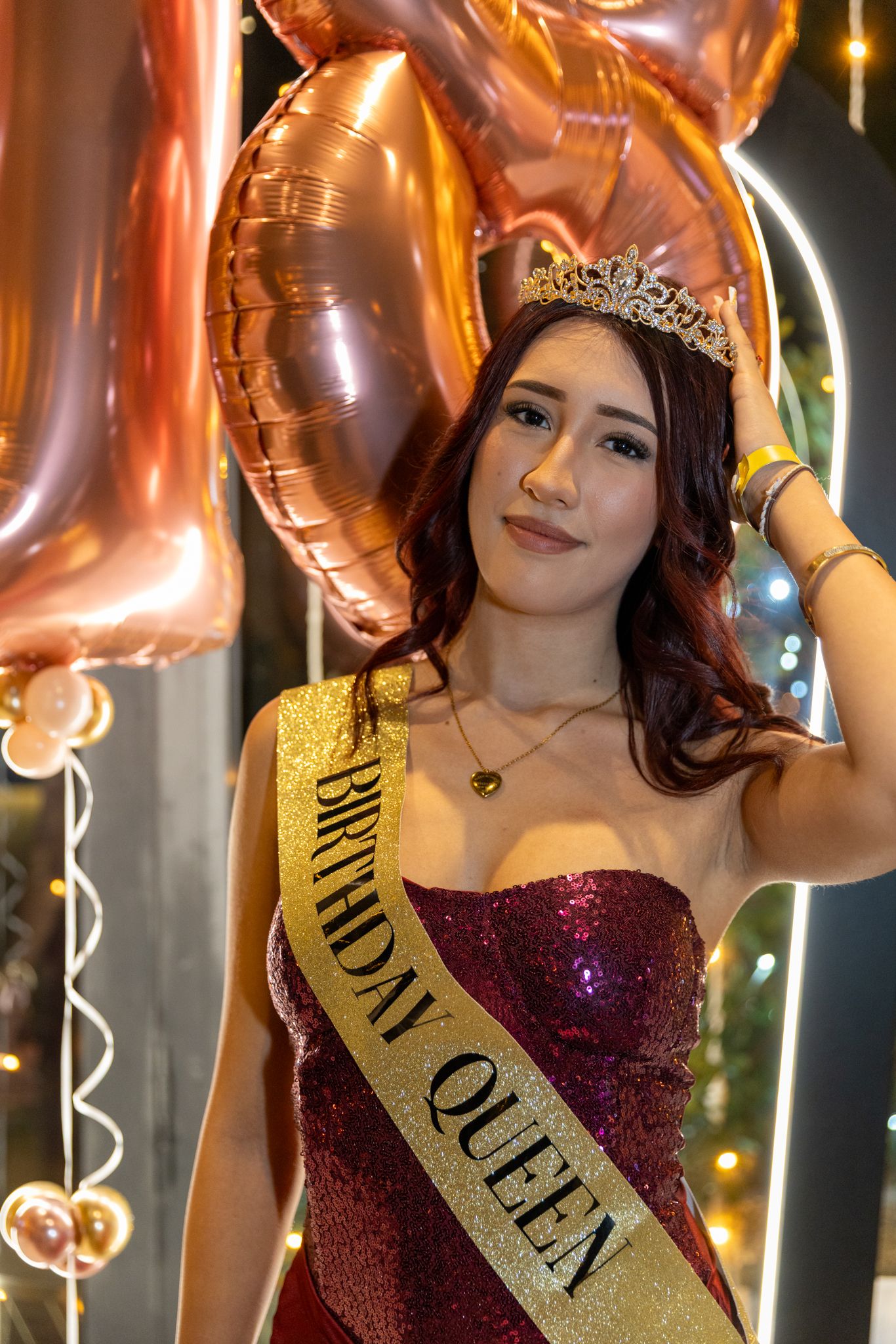 A person wearing a sparkly red dress, crown and gold sash with 'Birthday Queen' text poses with metallic balloons.