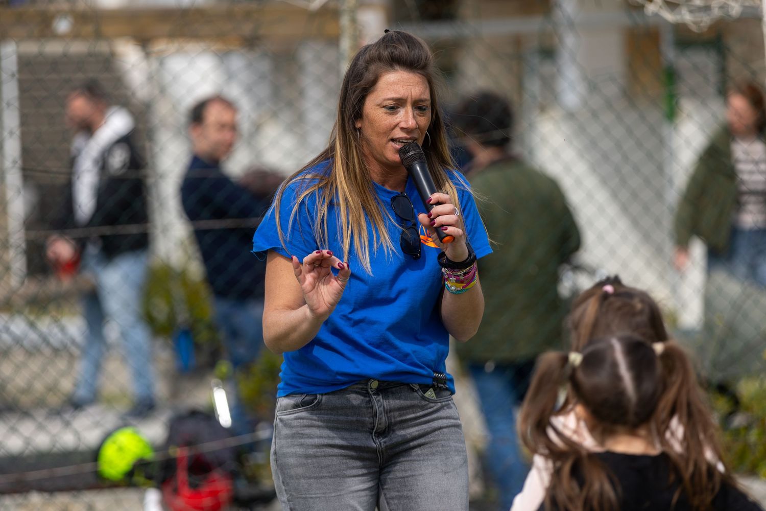 Woman in blue shirt speaking into microphone outdoors at a public event with crowd in background.