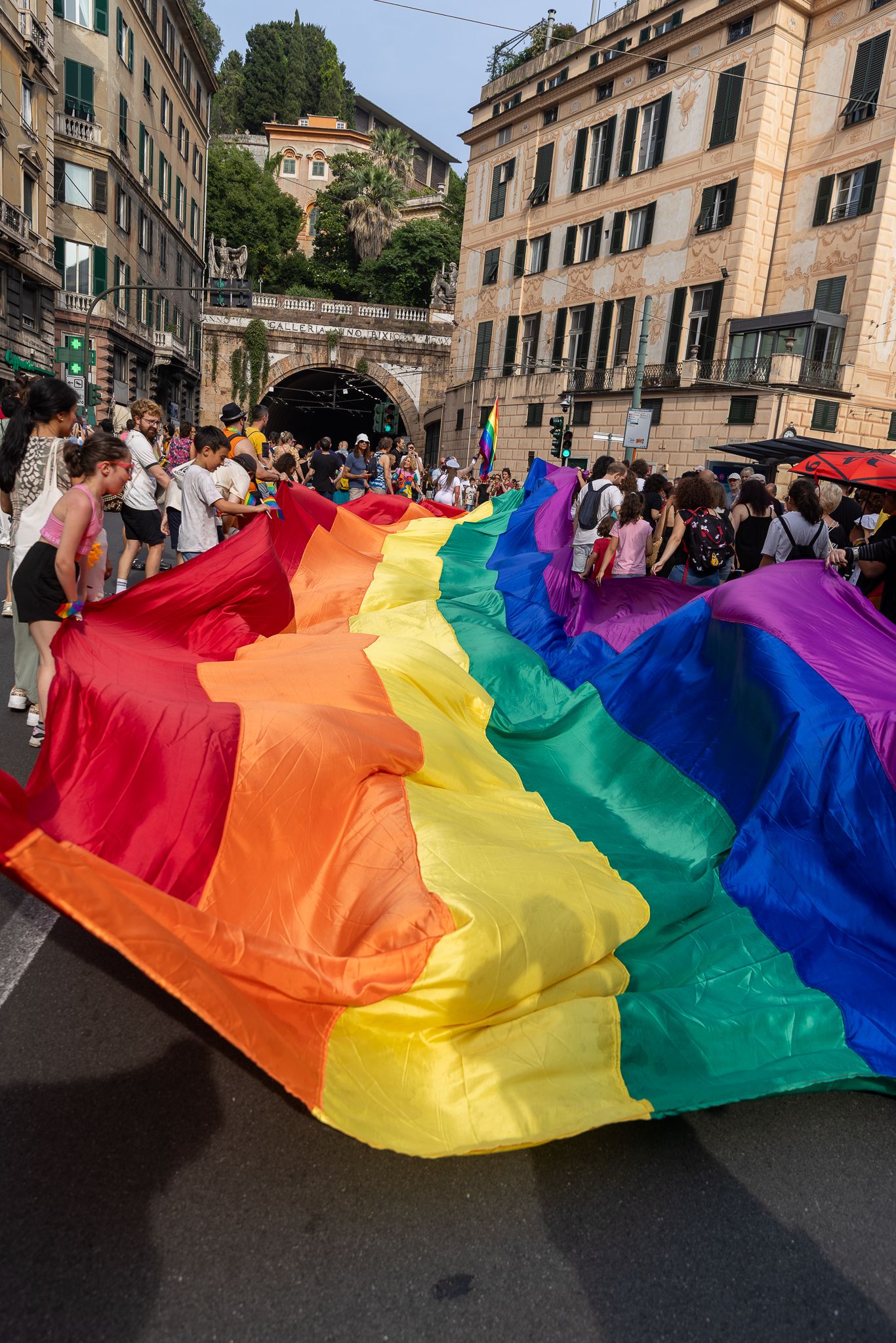 Large rainbow pride flag being carried through historic European city streets.