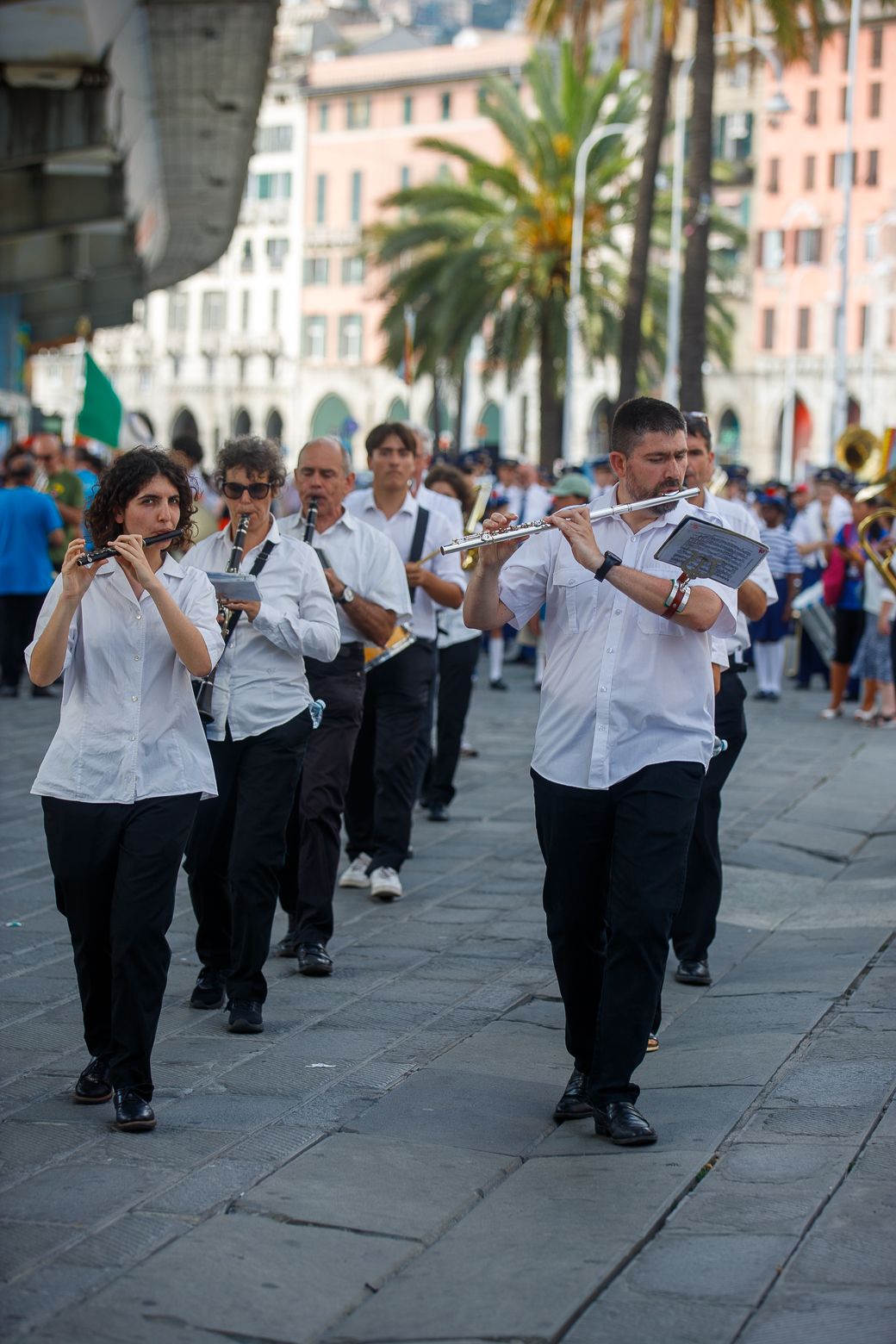 Marching band in white uniforms performing while walking through a palm-lined street.