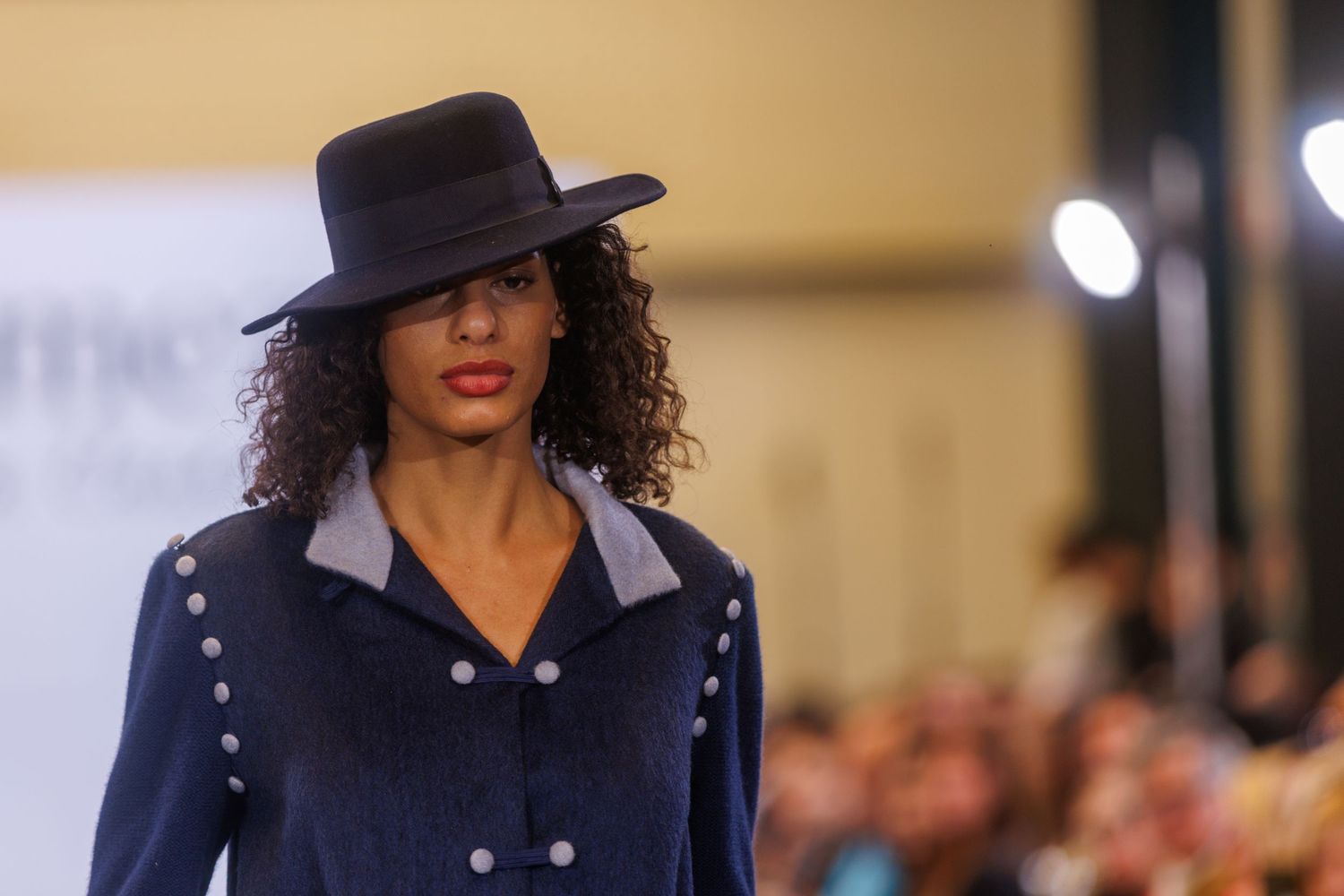 Model walks runway wearing navy blue military-inspired jacket and hat.