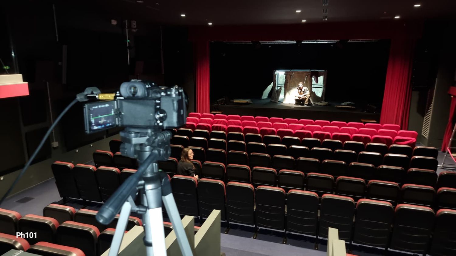 A professional camera on tripod set up in an empty red-seated cinema theater with a screen displaying an image.