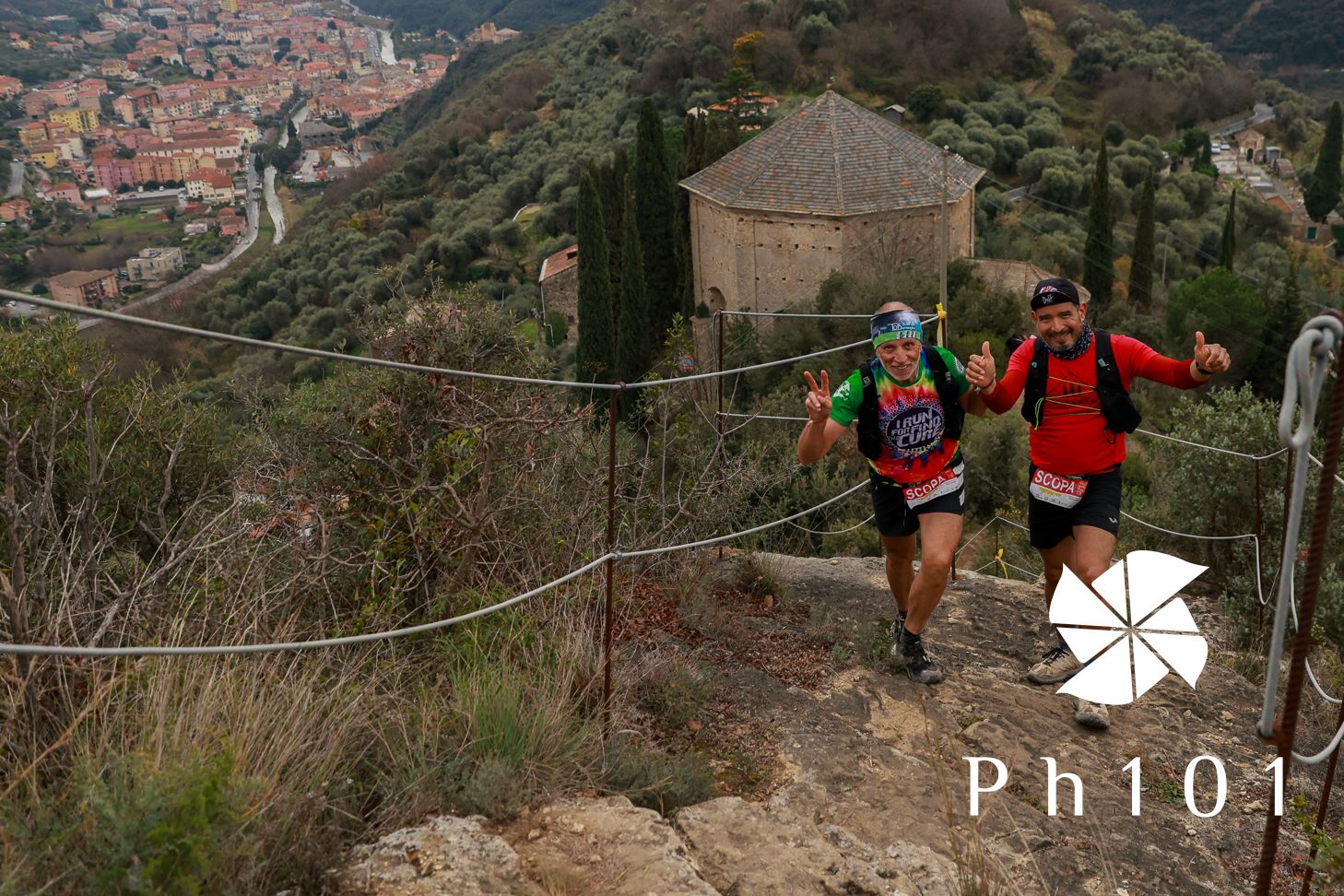 Trail runners navigate a steep mountain path with ropes while descending towards a distant town in the valley.